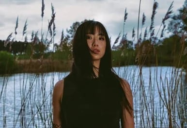 Woman with long dark hair standing by a lake with tall reeds in the background on a cloudy day