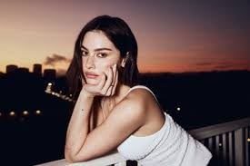 Woman with dark hair in white tank top posing at sunset with city lights in background