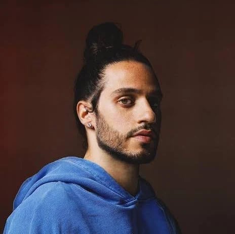 Portrait of a man with dark hair in a bun, wearing a blue hoodie, against a brown background
