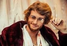 Man with reddish-brown curly hair wearing a red and black plaid jacket over a white shirt, smiling at camera