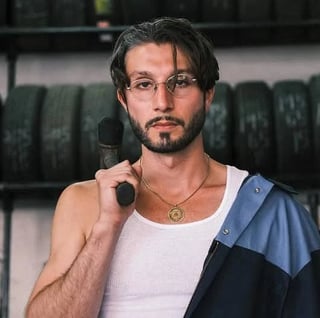 Man with dark hair and glasses wearing white tank top and blue jacket, holding headphones around neck