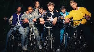 Group of eight men posing together on motorcycles and bicycles against a dark background