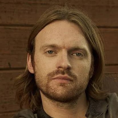 Man with shoulder-length brown hair and beard looking at camera against brick wall background
