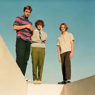 Three young men standing on a white platform against a blue sky, wearing casual 1970s-style clothing