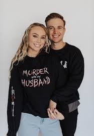 A couple poses together smiling at the camera, both wearing black hoodies against a white background