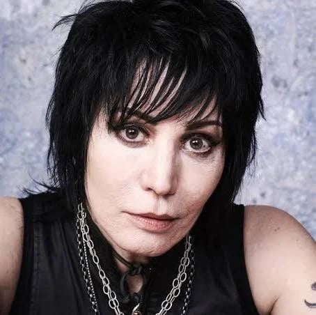 Person with black shaggy hair and dark makeup wearing a black sleeveless shirt and silver chain necklace against a textured gray background