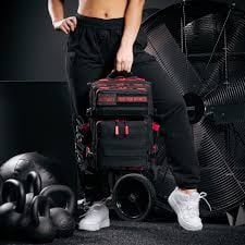 Person holding black gym backpack with red accents, surrounded by dumbbells, shoes, and workout gear