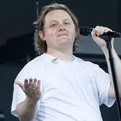 Man with shoulder-length hair performing on stage with microphone, wearing white t-shirt