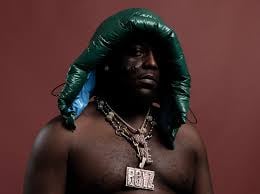 Man with long black hair and blue accents wearing multiple gold chains against a burgundy background