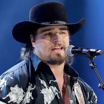 Male performer singing into microphone wearing black cowboy hat and ornate black and white jacket against blue background
