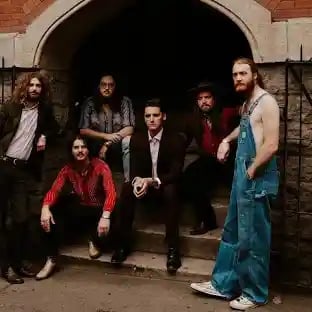 Group of seven people with long hair posing together in front of a brick archway entrance