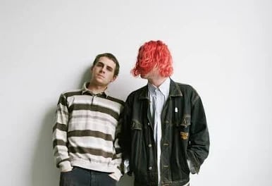 Two young people standing against white wall. Left person wears striped sweater and black pants. Right person has bright pink hair and wears dark jacket over white shirt.
