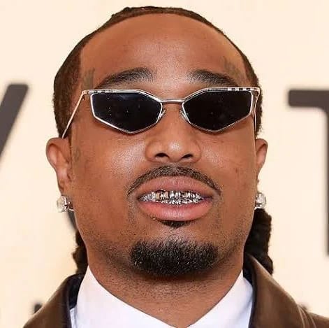 Man wearing rectangular sunglasses with dark lenses, diamond grillz, and hoop earrings against a beige background