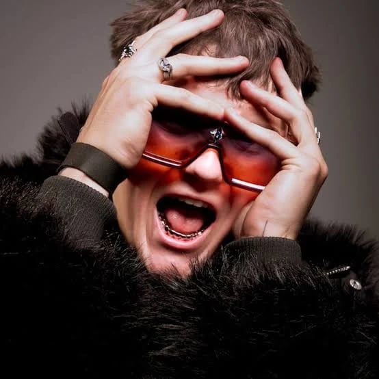 Person with mouth open wearing red-tinted sunglasses and black fur, hands framing face against gray background