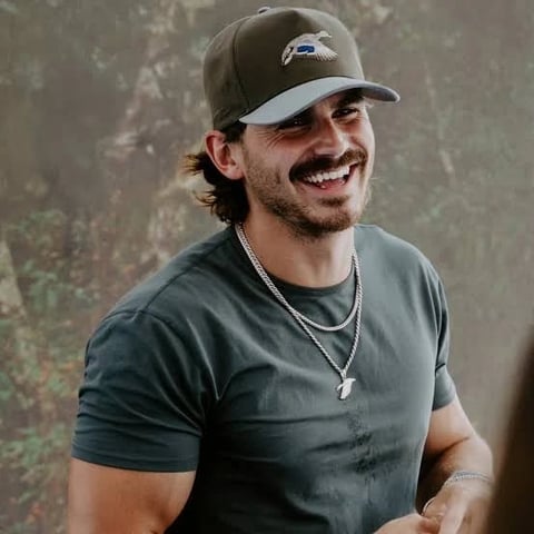 Man smiling at camera wearing gray cap and dark t-shirt with silver chain necklace