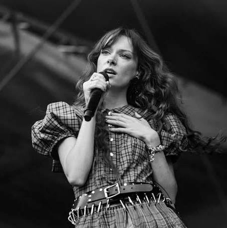 Black and white photo of a female performer singing into a microphone while wearing a plaid shirt and studded belt