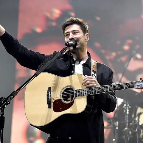 Male musician performing on stage with acoustic guitar and microphone, raising fist, against blurred backdrop with red lighting