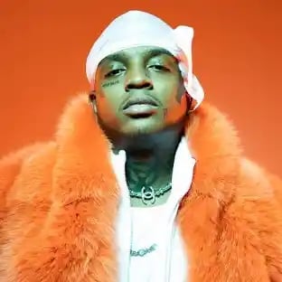 Man wearing white head wrap and orange fur coat against orange background, looking at camera