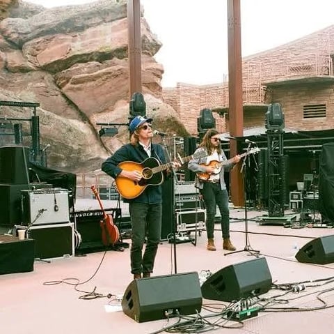 Two musicians performing on stage with guitars against a backdrop of canyon cliffs and green equipment, surrounded by microphones and sound gear