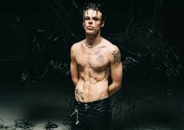 Shirtless muscular man with dark hair wearing black pants against a dark textured background