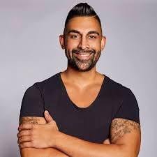 Man with dark hair and beard wearing a black v-neck t-shirt, smiling with arms crossed against a gray background