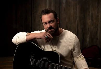 Bearded man in cream sweater holding a black acoustic guitar, posed thoughtfully against a dark wooden background.