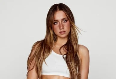Portrait of a woman with long brown hair wearing a white sleeveless crop top against a neutral gray background