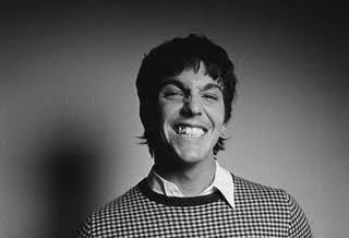 Black and white portrait of a smiling man with dark curly hair wearing a checkered shirt