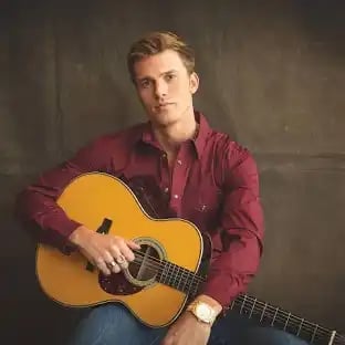 Male musician in burgundy shirt holding an acoustic guitar against a brown background
